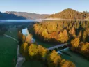 Luchtfoto van een rivier met brug door een herfstbos, met mist en bergen op de achtergrond.