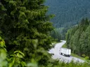 Vrachtwagen op een kronkelende bergweg door een dennenbos met bergen op de achtergrond.
