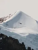 Besneeuwde bergtop met gletsjer en een kleine helikopter in de lucht.
