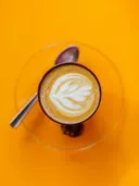 Close-up van een cappuccino met latte-art in een glazen kopje op een oranje tafel, met een lepel ernaast.