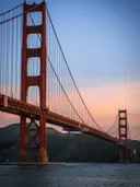 Verticale opname van de Golden Gate Bridge van onderaf met roze en oranje avondlucht en glooiende heuvels op de achtergrond.