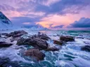 Golven die breken op rotsen aan de kust bij zonsondergang met besneeuwde bergen.