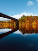 Brug over een rivier met spiegelend water en bomen in herfstkleuren.