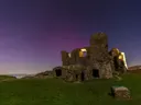 Verlichte kasteelruïne op een heuvel onder een sterrenhemel met paarse gloed, met de kust op de achtergrond