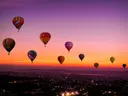 Meerdere kleurrijke heteluchtballonnen in de lucht bij zonsondergang boven een stad.