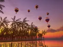 Meerdere kleurrijke luchtballonnen boven een rij palmbomen bij zonsondergang, met water op de voorgrond.