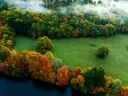 Luchtfoto van een groen weiland omringd door bos in herfstkleuren met mist.