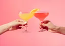 Foto van twee handen met cocktailglazen tegen een roze achtergrond