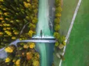 Luchtfoto van een rivier met waterval en brug, omgeven door herfstbos en een groen veld.