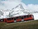Rode trein rijdt over een groene helling met een besneeuwde berg op de achtergrond.