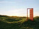 Surrealistisch beeld van een losstaande rode deur in een grasveld bij zonsondergang.