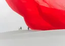 Surrealistisch beeld van een grote rode stofdoek zwevend boven witte duinen met drie wandelaars
