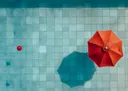 Bovenaanzicht van een rode parasol en rode bal op blauwgroene zwembadtegels