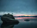 Bonsai Rock met kleine boompjes op een rots in kalm meerwater bij zonsondergang, met bergen op de achtergrond.