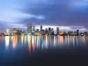 Stadssilhouet met verlichte wolkenkrabbers dat weerspiegelt in rustig water bij avondschemering.