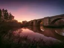 Historische stenen boogbrug over een rustige rivier bij zonsondergang, met weerspiegeling in het water.