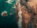 Luchtfoto van een rotsachtige kust met turquoise zee en golven op zandstrand.