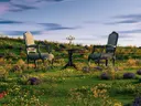 Twee antieke stoelen met een kandelaar op een tafel, midden in een heuvelachtig veld vol wilde bloemen bij schemering.