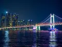 Verlichte hangbrug naast een stadsskyline bij nacht, weerspiegeld in het water met een boot ervoor.