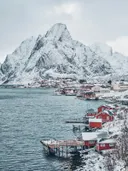 Rode vissershuisjes aan een fjord met een besneeuwde bergtop op de achtergrond.