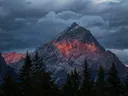 Bergtop die rood oplicht door alpengloed onder een bewolkte lucht met dennenbomen op de voorgrond.