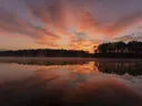 Een spiegelend meer omzoomd door dennenbos onder een felgekleurde avondlucht met wolkenstrepen.